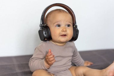 A baby in large headphones sits on a bed and listens attentively. The soft indoor lighting adds a peaceful and focused mood.