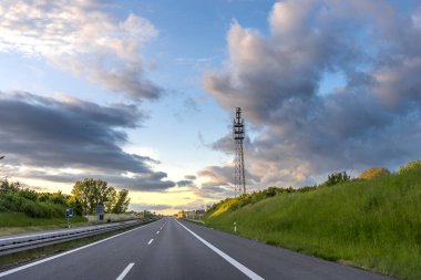 Boş bir otoyol, açık bir arazide uzanır. Yumuşak bulutlu, altın bir akşam gökyüzünün altında..