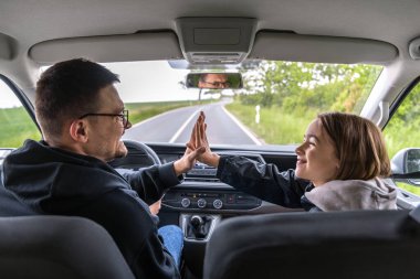 Bir adam ve bir kız birlikte araba sürerken avuç içlerine dokunuyorlar. Gülümsemeleri ve göz teması huzurlu bir bahar yolculuğunda sıcak bir bağı yansıtır..