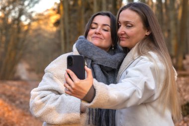Ormanda telefonlarıyla selfie çeken iki arkadaş. Doğadaki arkadaşlar. Yüksek kalite fotoğraf. İlkbahar, sonbahar mevsimi. Ormanda bir yürüyüş, doğada..