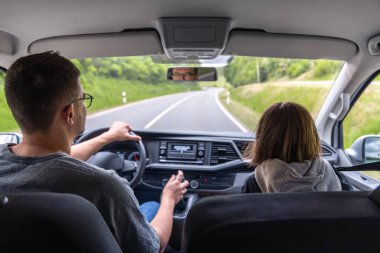 Kapüşonlu bir kız ön koltukta oturuyor ve ileriyi işaret ediyor. Islak bir orman yolunda yol gösteriyor. Yeşil bahar manzarası tazelik ve derinlik ekler..
