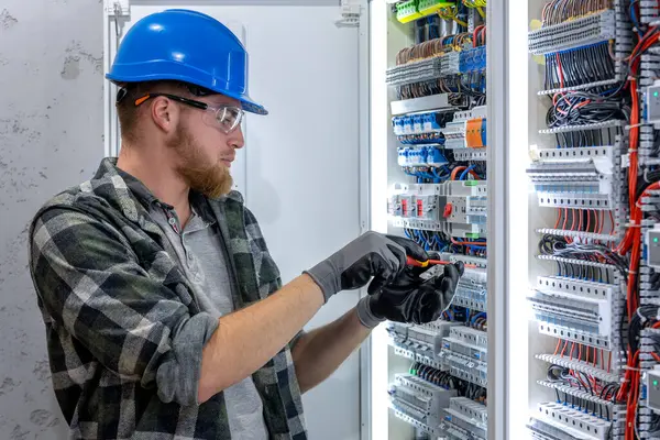 Güvenlik teçhizatı olan bir teknisyen güç panelinin önünde tablet tutuyor, odaklanarak yan bakıyor. Endüstriyel bir ortamda teknik olarak hazır olma sahnesi.