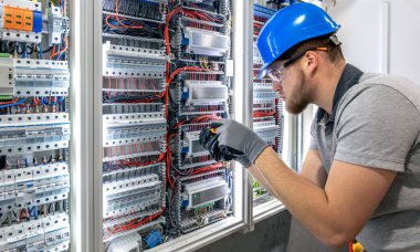 Bir elektrik teknisyeni, elektrik panelinin içine bileşenler yerleştiriyor. Yapay ışıklandırma, bir kişi, kapalı mekan ayarları, hassas teknik işler..