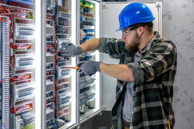 Koruyucu teçhizatlı elektrikçi elinde bir tornavida tutuyor ve kablolarla ve teknik donanımlarla çevrili bir panelin içindeki elektrik bağlantılarını dikkatle inceliyor..
