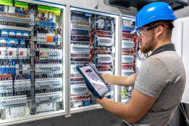 Sert şapkalı bir adam santrali elle incelerken tablet kullanır. Yapay ışıklandırma ve teknik hassasiyetle dijital tanı ve pratik eylem dengesi.