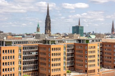 Kırmızı tuğlalı binalar ve tarihi kilise kulelerinin yer aldığı panoramik bir şehir manzarası. Eski ve modern mimarinin çarpıcı kontrastı.