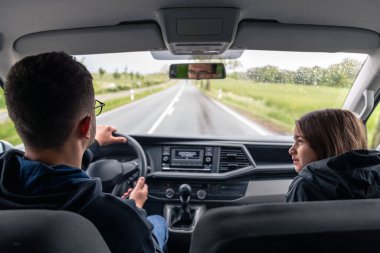 Araba sürerken yanında ceketli bir kız otururken pencereden dışarı bakan bir adam. Dışarısı yağmurlu, sakin bir atmosfer..