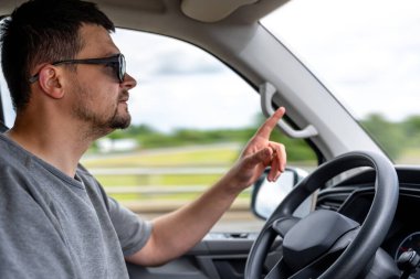 Güneş gözlüklü bir adam işaret parmağıyla arabayı ileri doğru sürüyordu. Kabinin içinden yan görüntü, jest ve yoğunluğu yakalıyor..