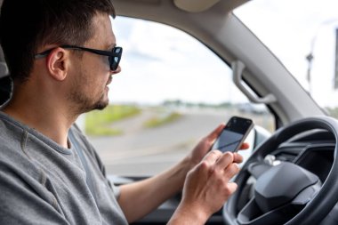 Direksiyonun arkasında akıllı telefon tutan güneş gözlüklü bir adam mola sırasında navigasyon uygulaması ile etkileşime geçiyor. Sürücü ve gösterge paneline odaklanmayı temizle.