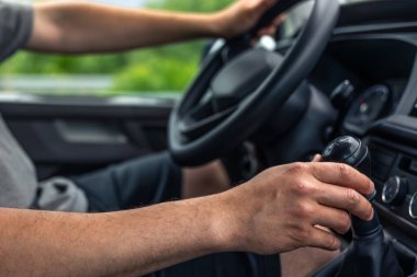 Araba sürerken vites değiştiren bir adamın yakın çekimi. İki el de direksiyonda ve vites kolunda görünür..