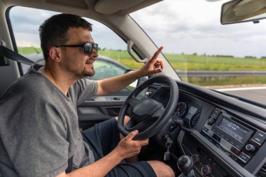Güneş gözlüğü takan, araba süren ve parmağıyla hareket eden bir adam. Arkaplanda kırsal yol ve araç.
