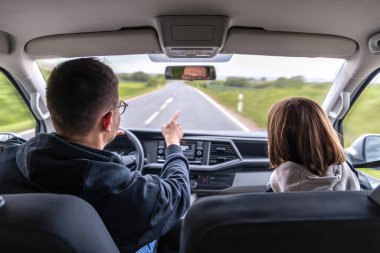 Yolcu koltuğundaki bir kız yeşil orman yolunda giderken yönü işaret ediyor. Arabanın içi bir güven ve sükunet hissini yansıtıyor..