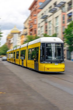 Şehir caddesi boyunca ilerleyen sarı bir tramvayın dikey görüntüsü. Hareket bulanıklığı, karışık mimarinin arka planına karşı hız duygusunu arttırır..