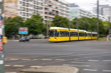 Uzun sarı bir tramvay hareket bulanıklığı olan geniş bir kavşakta hareket ediyor. Sahne şehir ölçeğini ve toplu taşımanın hızını aktarıyor.