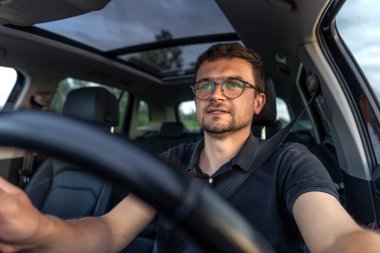 Modern bir araba sürerken gözlüklü bir adam hafifçe gülümsüyor. Günışığı ve iç ortam rahatlığı, odaklanmayı ve sakinliği yansıtıyor..