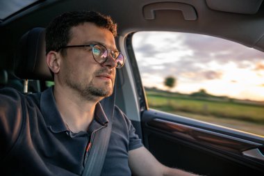 Gün batımında araba kullanan gözlüklü bir adamın yakın çekimi. Arka planda hareket bulanıklığı var. Otoyolda hız ve hareketi vurguluyor..