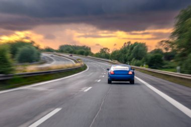 Mavi bir araba yüksek hızda virajlı bir otoyolda hızla ilerliyor, dinamik bulanıklık etkisi var. Parlak bir günbatımı sıcak ve dramatik bir hava yaratır..