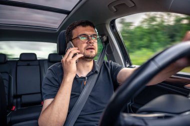 Araba kullanırken telefonla konuşan bir adam modern bir aracın içinde dikkati dağılmış davranışları ve yol güvenliği endişelerini vurguluyor..