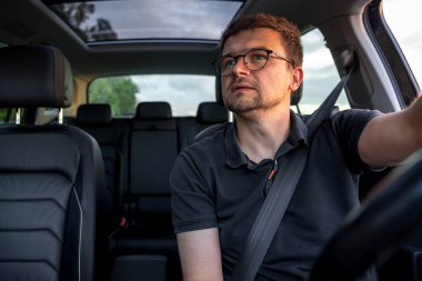 Siyah tişörtlü konsantre bir adam akşam saatlerinde araba kullanır, yan tarafa bakar, arabanın içindeki yumuşak ışık düşünceli bir ruh hali ekler..