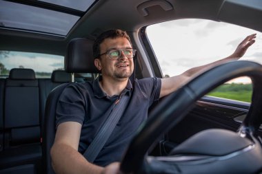 Pencereden elini kolunu sallayarak çıkan bir adam, yaz gezintisi sırasında gülümsüyor. Doğal ışık ve rahat duruş özgürlüğü ve mutluluğu ifade eder..