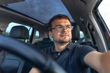 Bir adam araba sürerken hafifçe gülümser, yumuşak gün ışığıyla arabanın rahatlığını ve iç açıklığını arttırır..