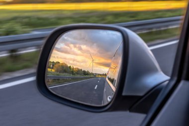 Bir rüzgar türbini altın gün batımında araba yan aynasına yansıyarak sürdürülebilir enerjiyi hareket özgürlüğü ve hareketlilikle harmanlıyor..