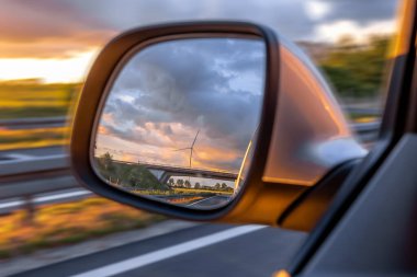 Sıcak günbatımı tonları, yolculuk, sürdürülebilirlik ve modern yenilenebilir enerji kavramlarına sahip bir rüzgar türbini araba aynasına yansıyor..