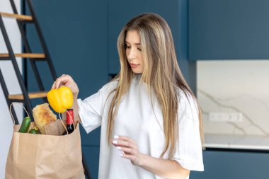 Modern bir mutfakta paketleri boşaltan kadın elinde parlak sarı bir dolma biber tutuyor. Sürdürülebilirliğe, tazeliğe ve dikkatli yemeğe odaklan.
