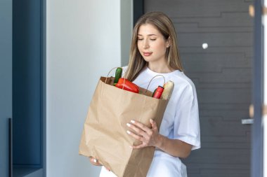 Ön kapının yanında elinde poşet tutan genç bir kadın. Doğal ışık, sakin ruh hali, çevre dostu alışveriş ve ev teslimatı kavramı.