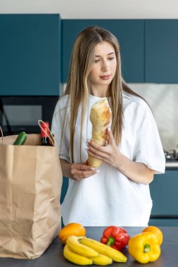 Modern bir mutfakta genç bir kadın kağıt bir torbadan meyve ve sebze paketleri açıyor, taze ürünler tezgahın üzerine serilmiş, canlı, çevreci bir sahnede..