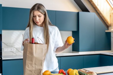 Modern bir mutfakta genç bir kadın poşetten portakal çıkarıyor. Parlak ışıklandırma, organik gıda ve sıfır atık yaşam tarzı.