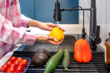 Kadın ellerinin temiz ve modern bir mutfakta akan suyun altında sarı biber yıkadığı yakın plan. Sessiz bir çevre dostu ritüeli..