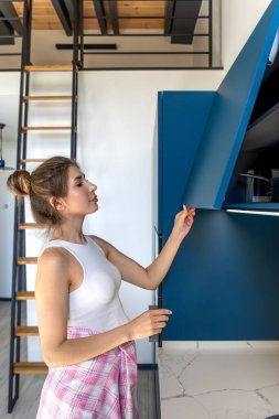 Genç bir kadın aydınlık bir odada mutfak dolabını açıyor. Ahşap ve mavi tonlu modern alan dengeli, minimalist bir iç mekan sunar.