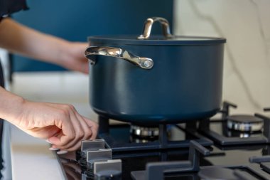Modern karanlık bir tencereyle gaz sobasını açan bir kadın elinin yakın çekimi. Ev yemeği ve minimalist mutfak hayatının başlangıcını gösteren detaylı bir çerçeve..