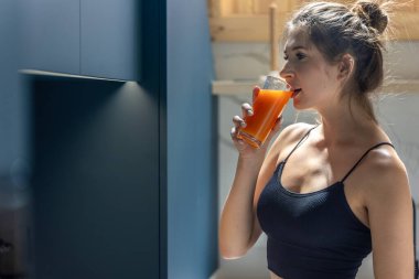 Spor kıyafetli güzel bir kadın sabah antrenmanından sonra sıcak bir mutfakta taze portakal suyu içer. Doğal ışık ve sağlıklı titreşim yaşam tarzını tanımlar.
