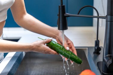 Su altında taze salatalık yıkayan ellerin yakın çekimi. Sağlıklı bir alışkanlık, temiz bir yaşam ve dikkatli bir yemek..