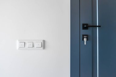 Parlak duvar, mavi kapının yanında siyah bir kilit ve anahtar, temiz çizgiler ve dengeli bir kontrastla, minimalist bir sahnede akıllı bir ev detayı ile değiştirilir..