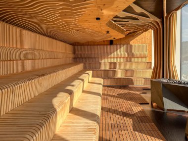 Parametrik banklar ve modern bir saunadaki dalgalı termotahta paneller, sıcak ışık ve grafik gölgeler, hiç insan olmadan heykel atmosferini şekillendiriyor..