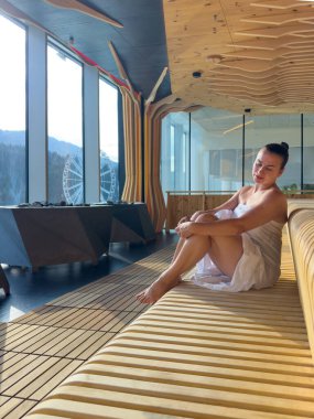 Havlu giymiş bir kadın dalgalı bir bankta oturuyor gözleri kapalı, yumuşak güneş ışığı ve panoramik pencereler sakin bir sağlık havası yaratıyor..