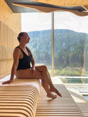 Kadın, dağlara ve göle bakan geniş bir pencerenin yanındaki tahta levhaların üzerinde modern bir saunada oturuyor. Doğal ışık ve kereste dokusu sakin ve sağlıklı bir ruh hali yaratır..