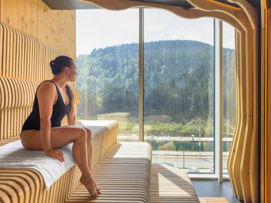 Kadın, dağlara ve göle bakan panoramik bir pencerenin yanında modern bir saunada oturuyor. Doğal kereste ve ışık sakin bir sağIık ortamı oluşturur..
