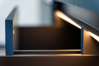 Çekmece rayları boyunca yumuşak LED aydınlatmalı alçak açı. Pürüzsüz hatlar, sıcak yansımalar ve mat mavi uçaklar minimalist zarafeti vurguluyor.