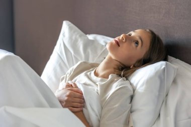 Beyaz bir yatakta yatıp yukarı bakan bir kız. Uyanık olduğumuz an gün ışığını, sakinliği ve minimalist ev estetiğini gösteriyor..