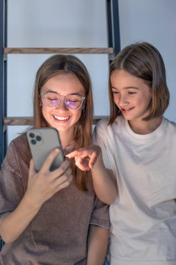 İki kız akıllı bir telefona bakar ve gülümser. Dijital iletişim ve gençlik tarzıyla evlerinde rahat ederler..