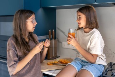 İki kız modern bir mutfakta konuşuyor, biri elinde bir bardak portakal suyu tutuyor, aile içinde sıcak bir an..