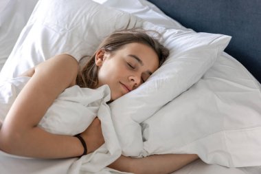 Beyaz bir battaniyeye sarılı, huzur içinde uyuyan genç bir kadın. Huzur, rahatlık ve uykusunun ahengi yayılıyor..