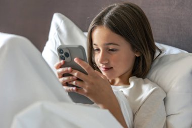 Kız beyaz bir yatakta yatıp akıllı telefondan mesaj atıyor. Parlak yatak ve temiz hatlar modern bir minimalist hissi yaratır..