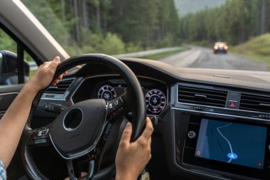 Direksiyonda elleri olan bir arabanın iç manzarası, ekranda GPS navigasyonu ve orman yolunda başka bir araba. Modern yaşam tarzı ve hareket ortamı.
