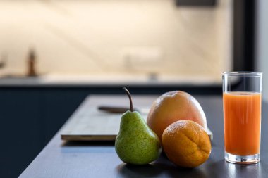 Meyvelerin yakın görüntüsü ve arkasında yumuşak mutfak ışığı olan koyu renkli bir tezgahın üzerinde uzun bir bardak portakal suyu. Lezzet, su ve enerji hakkında bir çerçeve.