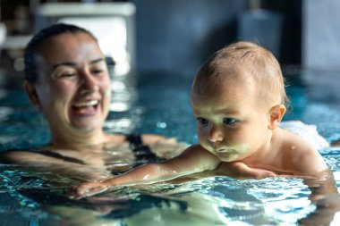 Anne sıcak bir spa havuzunda ona destek olurken bir bebek suda yüzüyor. Çocukların odaklanmış yüzü ve parlak yansımaları erken yüzme deneyimini yakalar..